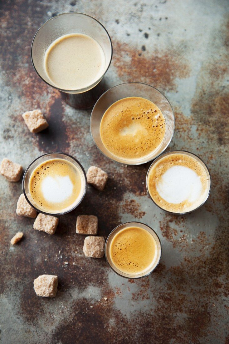Various glasses of coffee with sugar cubes on a metal surface