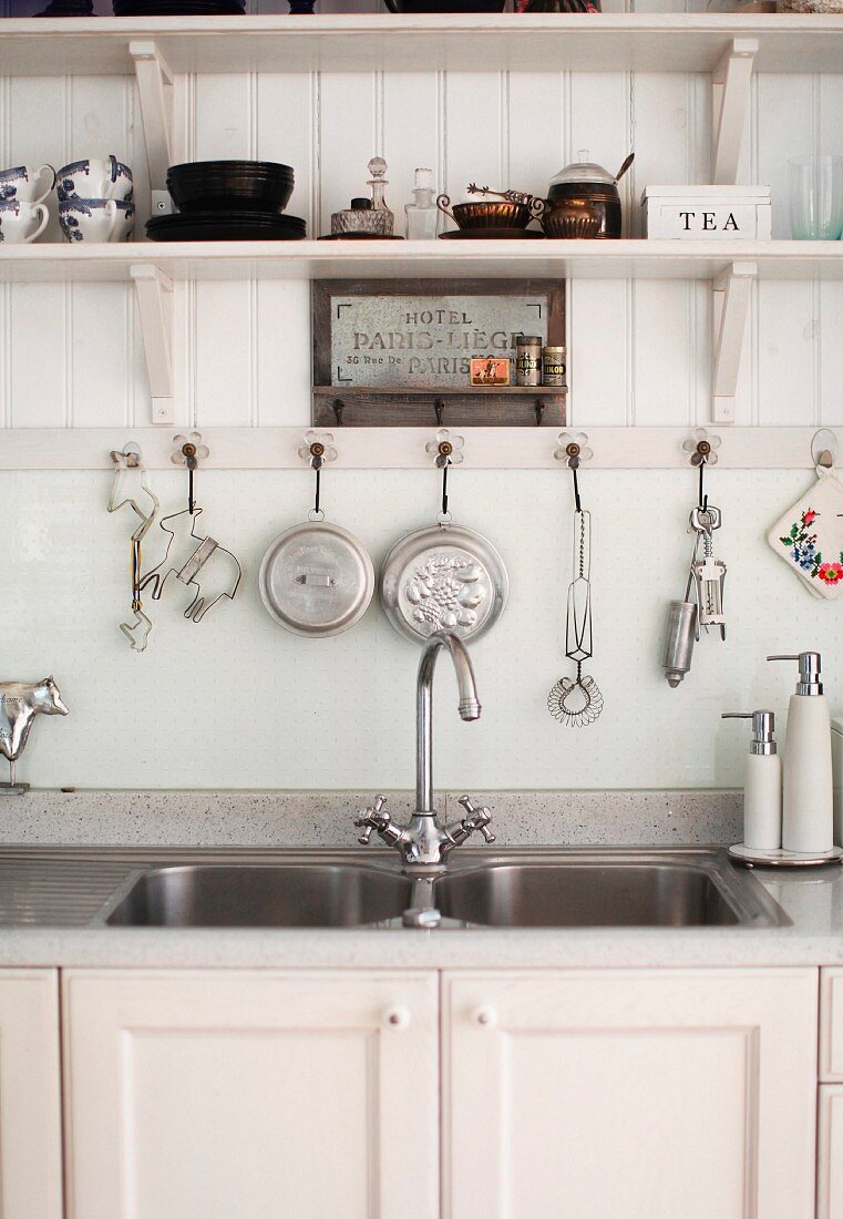 Twin sinks with white base cabinet below kitchen utensils hanging from hooks under wooden, wall-mounted shelf