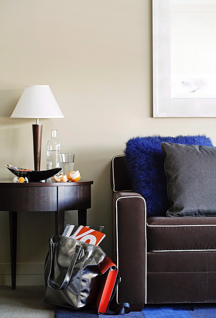 Round side table and table lamp next to dark grey armchair with white piping and shopping bag on floor