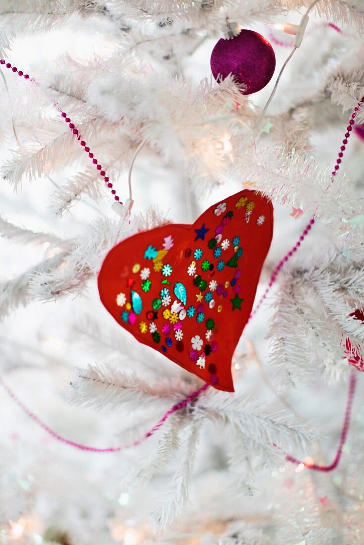 Red, hand-crafted love heart with colourful sequins and purple bauble on white, artificial Christmas tree