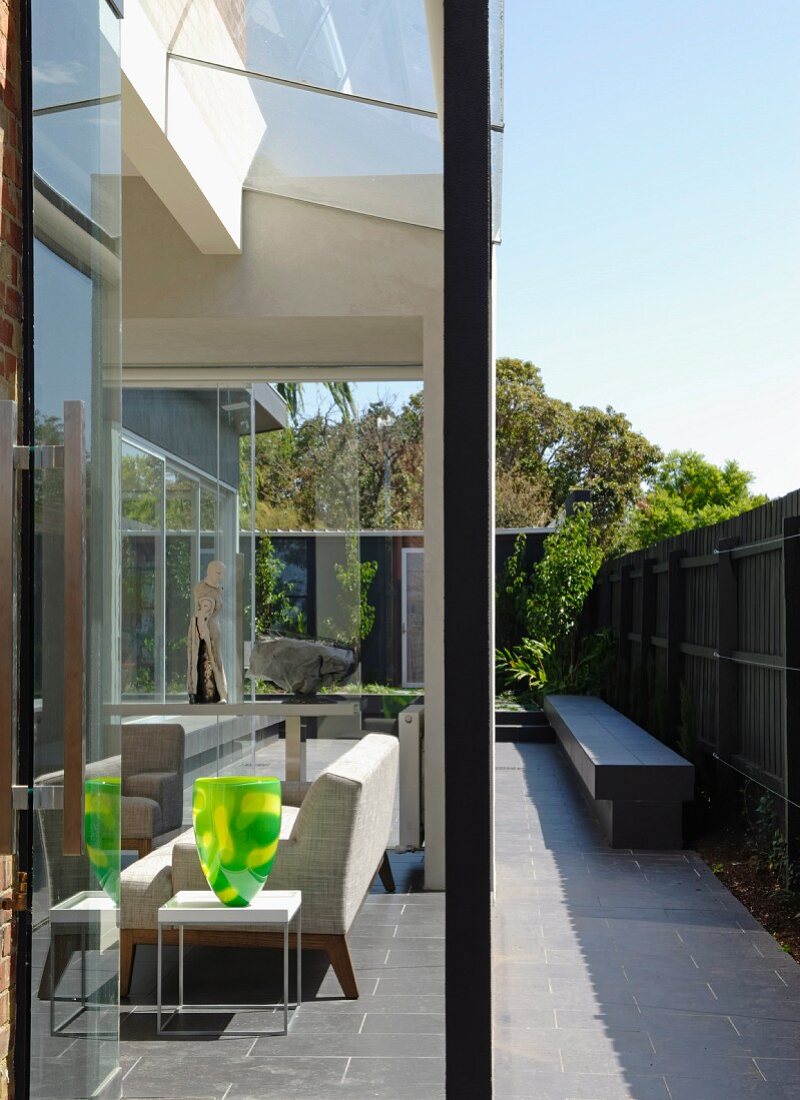 Glass walls create seamless transition between seating area in interior and narrow courtyard with stone bench against screen fence; decorative glass vase