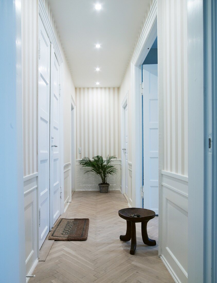 Narrow hallway with tall, white, traditional-style interior doors, striped wallpaper and rustic wooden stool on herringbone parquet floor