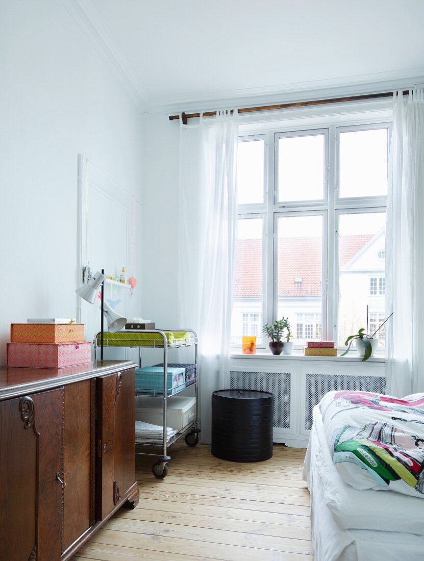 Old, dark wood sideboard and mobile shelves against wall in bright bedroom with wooden floorboards