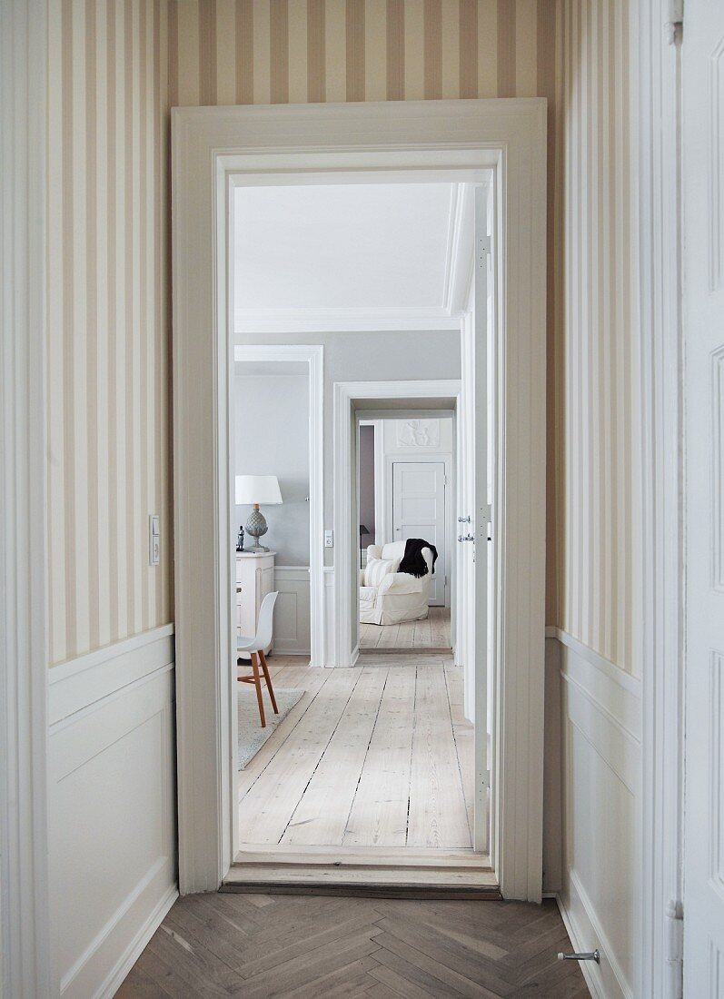 View from narrow hallway with striped wallpaper and herringbone parquet into living area with rustic wooden floor