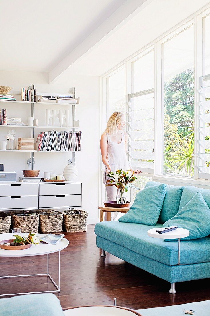 Sofa with light blue upholstery and white coffee table on dark parquet floor in bright, modern living room; woman standing at window in background