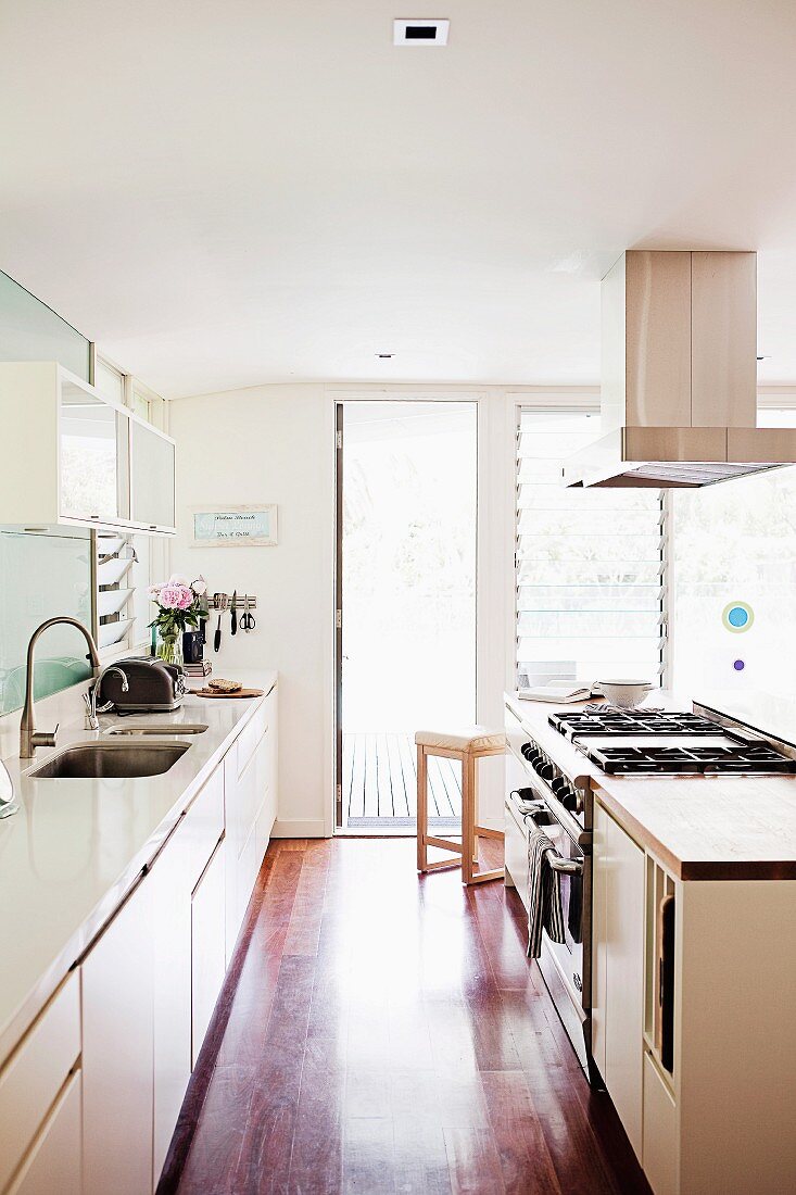 Parallel, white kitchen counters, extractor hood hanging from ceiling and glass, louvre terrace doors in background