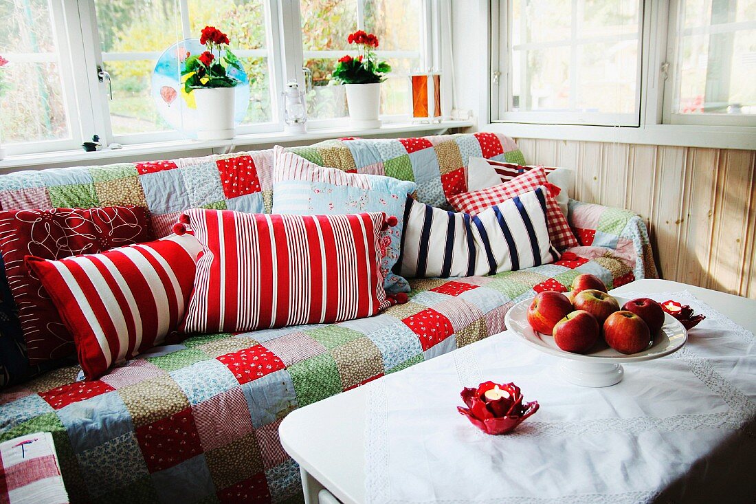 Comfortable corner in loggia - bowl of apples on white table in front of sofa with patchwork throw and patterned scatter cushions