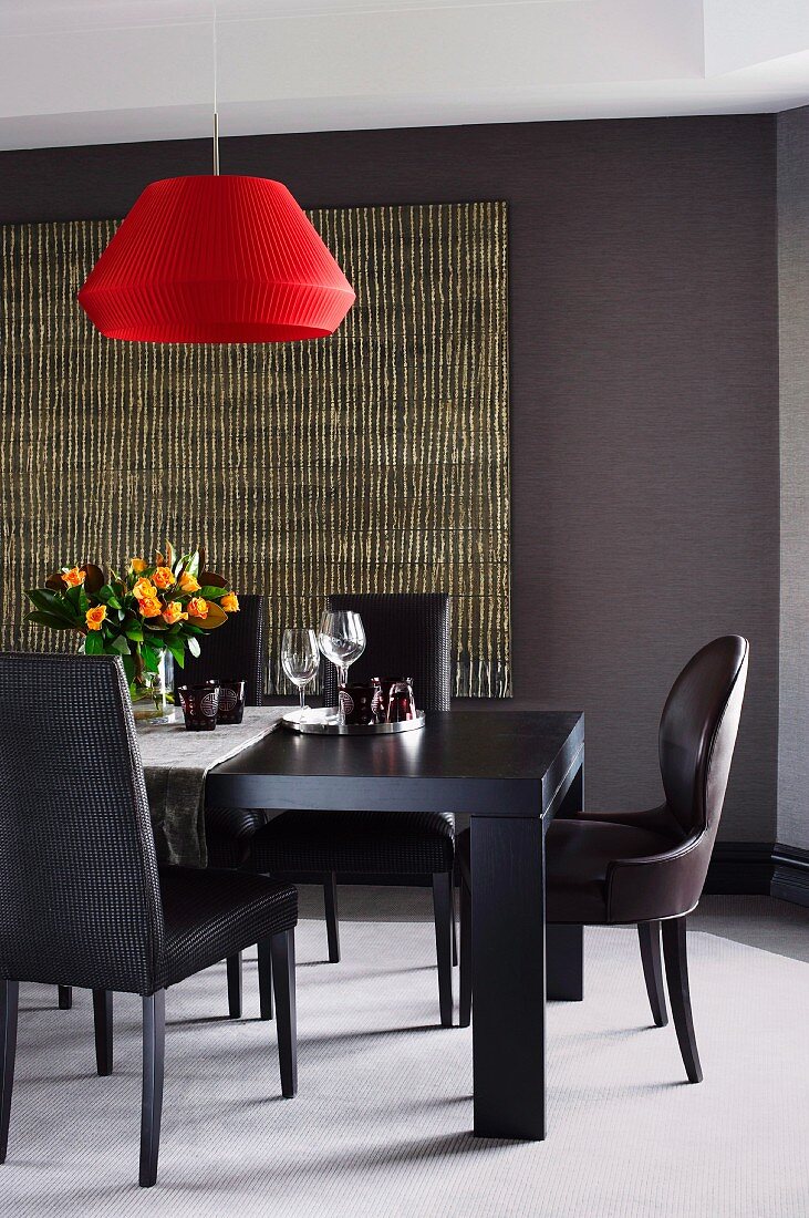 Elegantes Esszimmer, schwarze lederbezogene Stühle um massivem Esstisch, darüber Hängeleuchte mit rotem Schirm, an dunkler Wand Teppich aus Rattanstäben