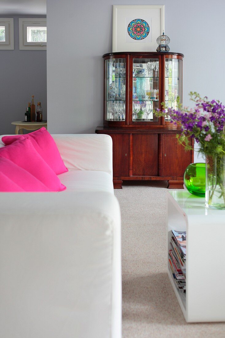 White sofa with pink scatter cushions and plastic side table; antique display cabinet against wall in background