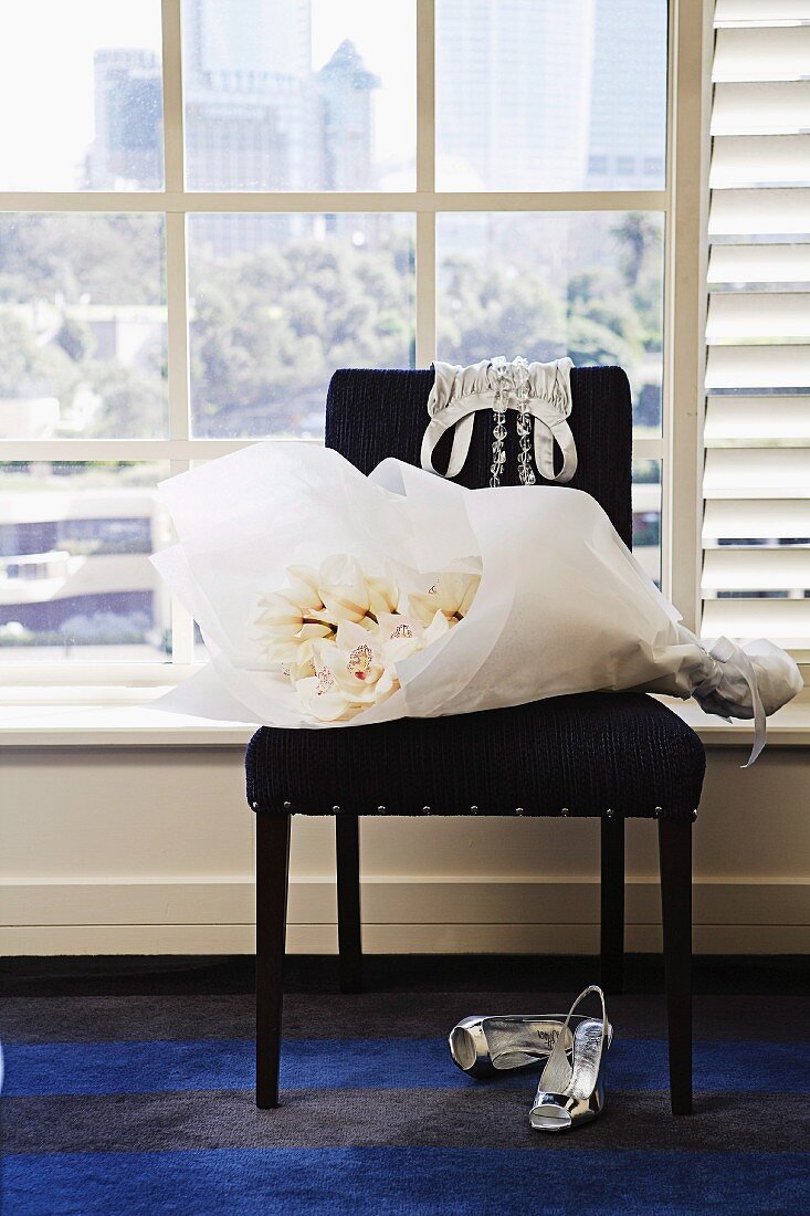 Weisser Blumenstrauss im Papier auf schwarzem, gepolstertem Stuhl und Damen Pumps auf Teppich vor Sprossenfenster