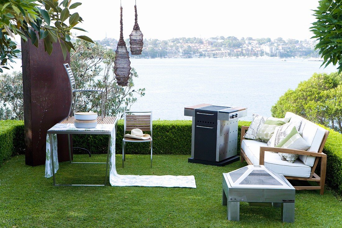 Idyllic seating area in garden with outdoor furniture, elegant barbecue, lanterns hanging from tree and sea view