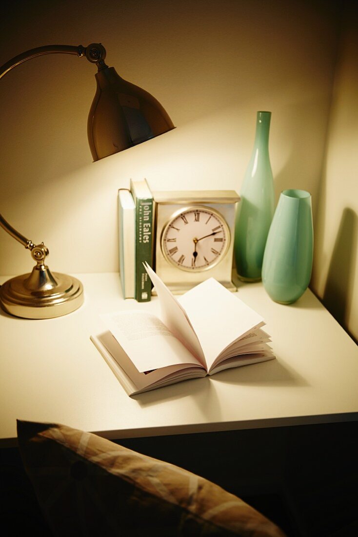 Open book below lamp on desk with clock and vases