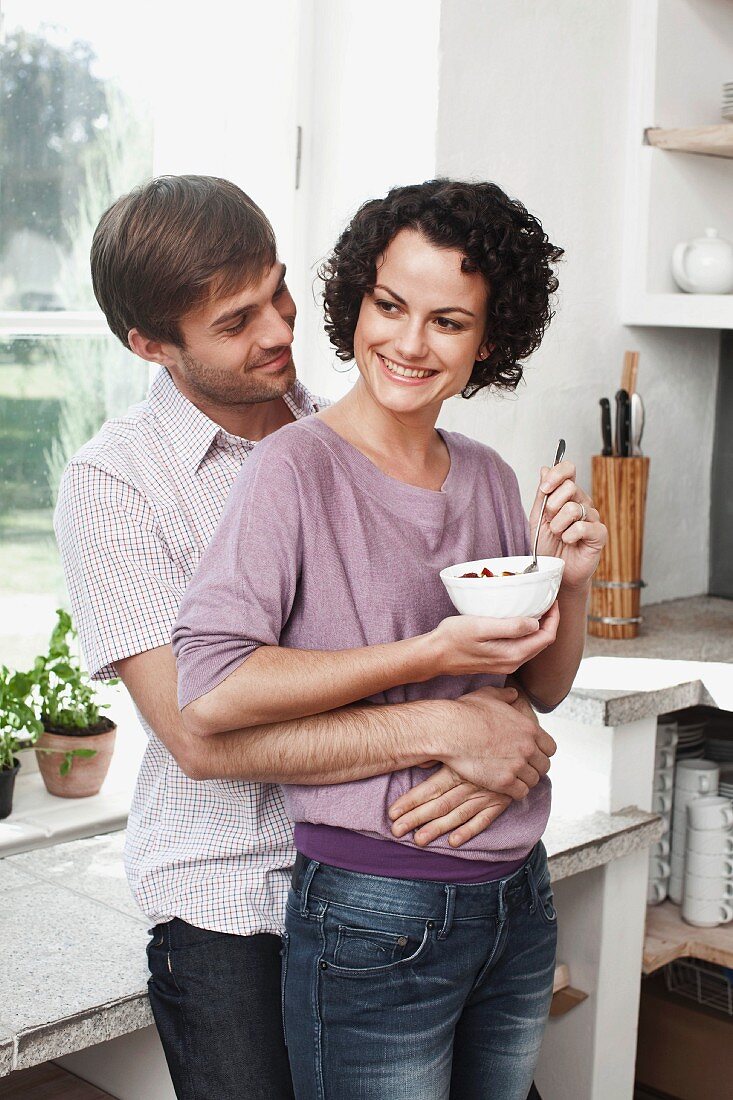 A couple embracing in the kitchen with the woman holding a muesli bowl