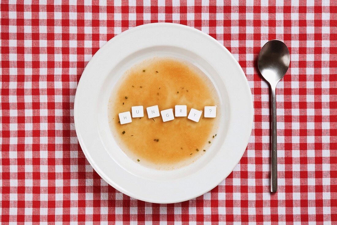 A bowl of soup on a checked tablecloth with computer keys floating in it spelling HUNGRY?