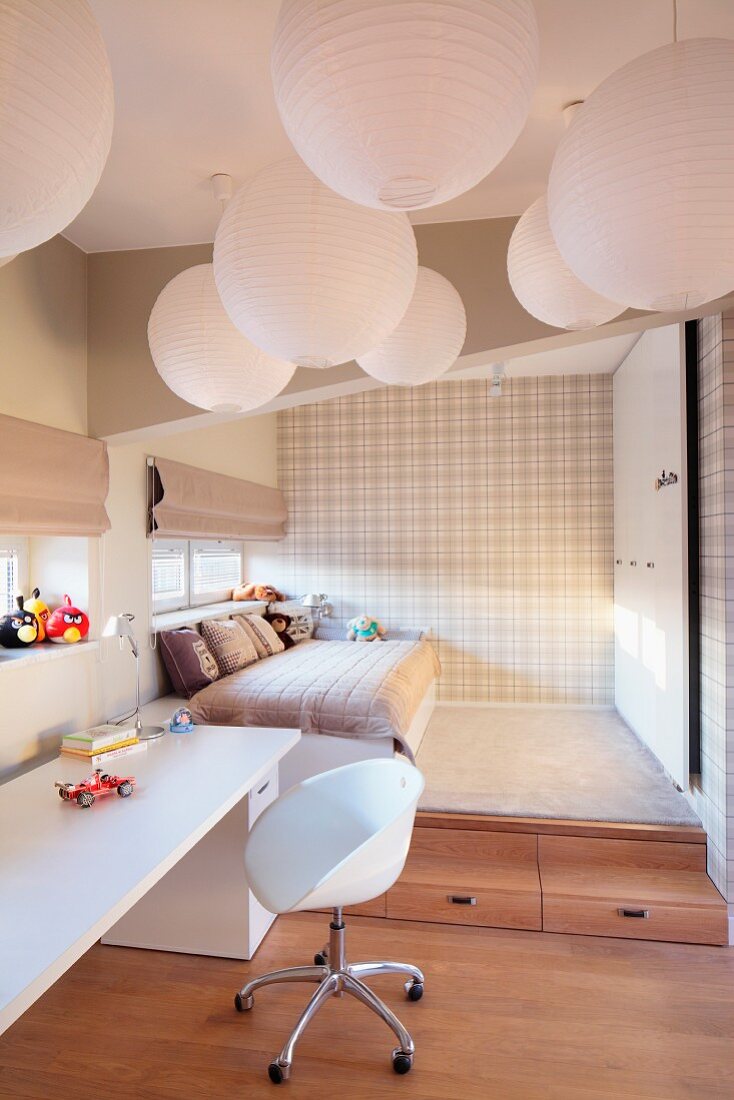 Multiple spherical pendant lamps made of white paper above swivel chair with white shell seat and desk; bed on platform in background in sleeping area with pale tartan wallpaper