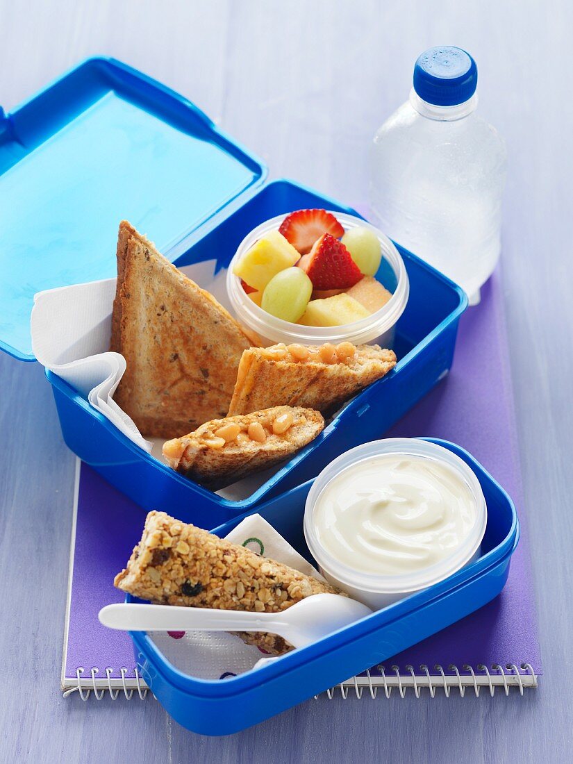 Lunchbox mit Sandwichtoasts mit Baked Beans, Obstsalat, Müsliriegel und Joghurt
