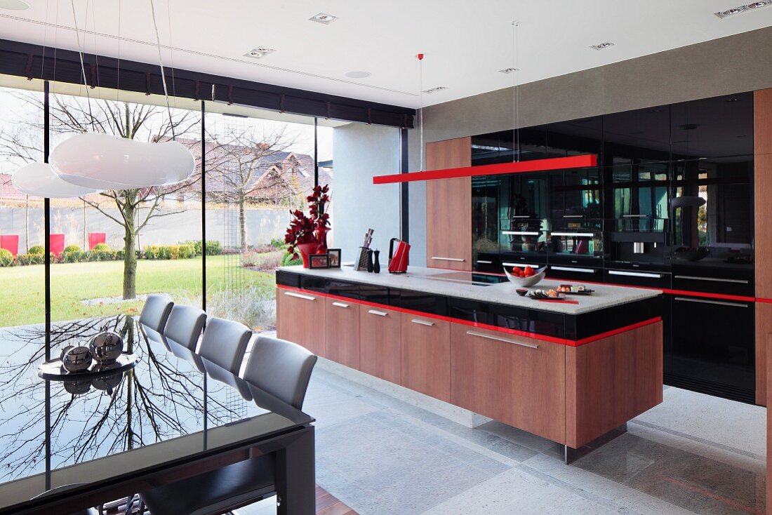 Dining area in front of kitchen area with island counter below delicate red pendant lamp and fitted cupboards with black fronts; glass wall with view into courtyard