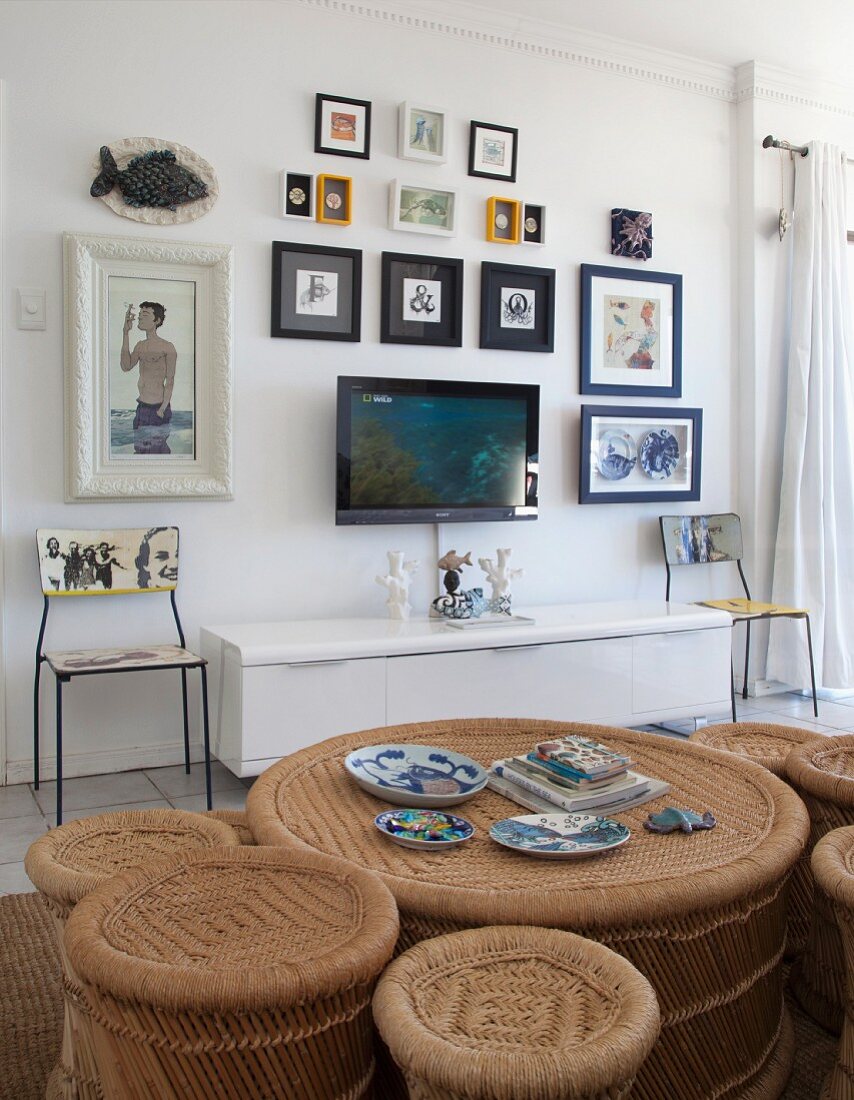 View across rattan coffee table with matching stools in various sizes to flatscreen TV and gallery of pictures on wall above white sideboard