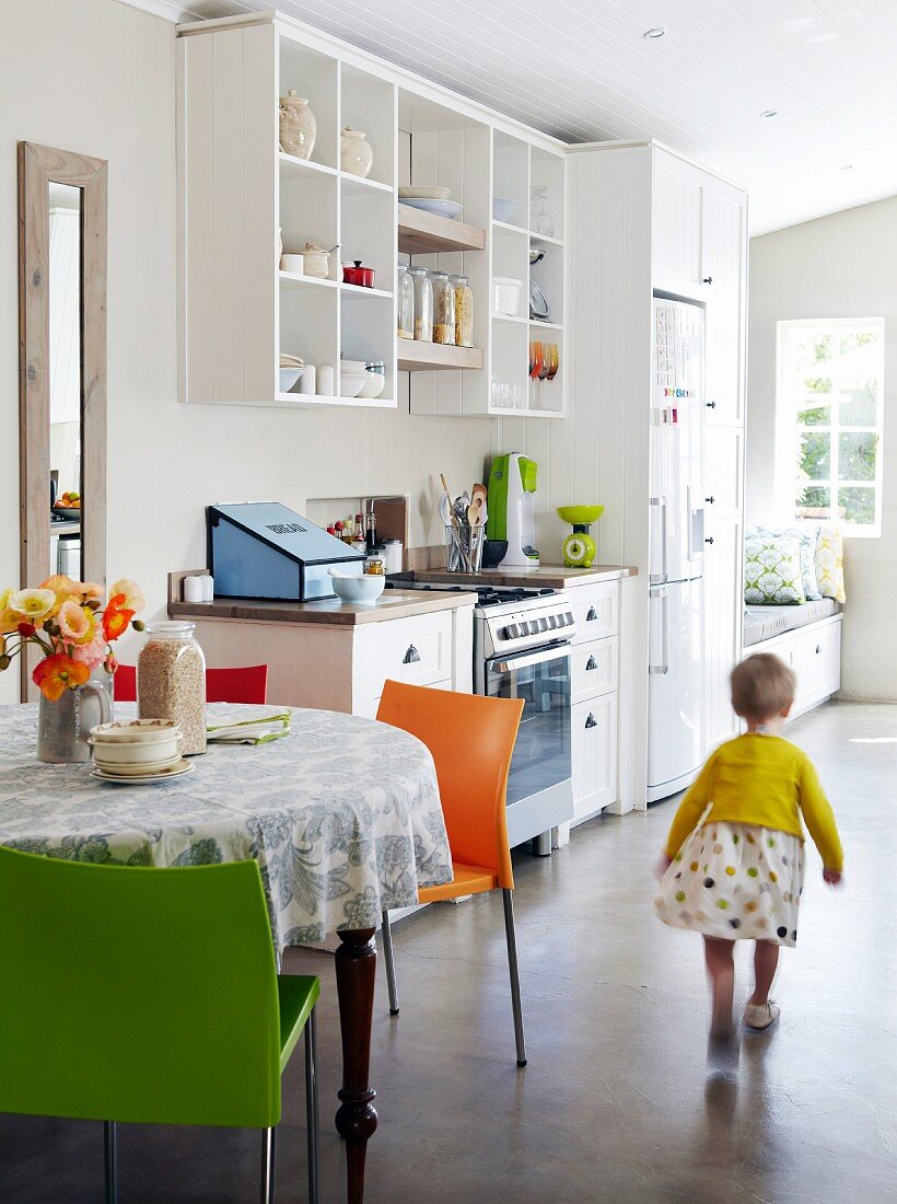 Kleines Mädchen in heller lichtdurchfluteter Küche mit pflegeleichtem Zementboden, offenem Küchenregal & rundem Esstisch mit farbigen Stühlen