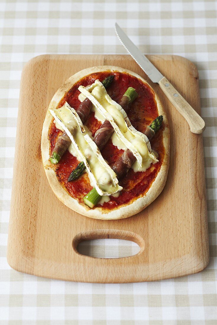 A mini pizza with green asparagus and brie on a wooden board