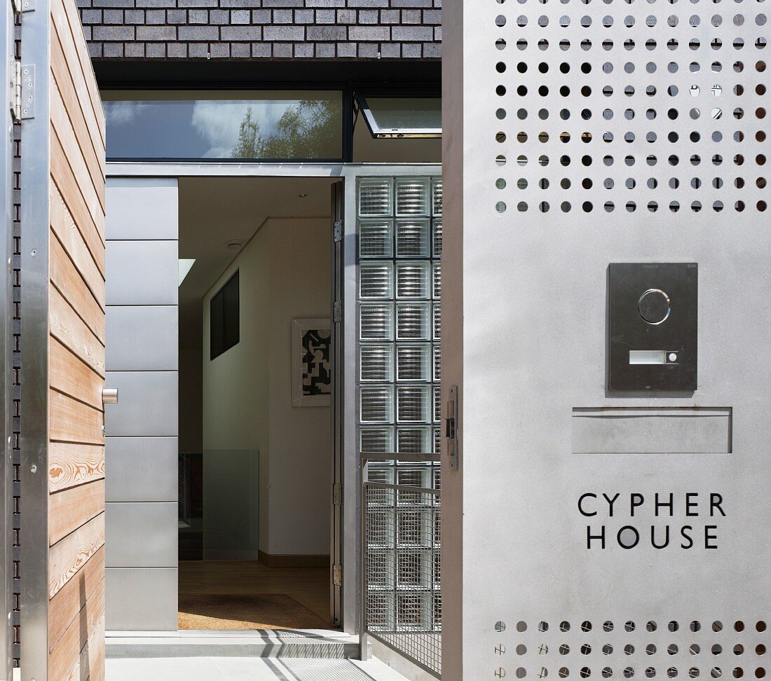 View through an open garden gate next to a buzzer on a perforated plate and an open front door