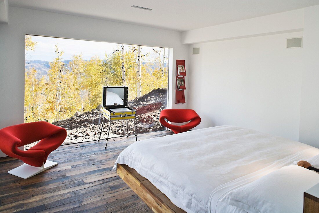 White bedroom with red designer armchairs in front of a large bank of windows with a view of the countryside