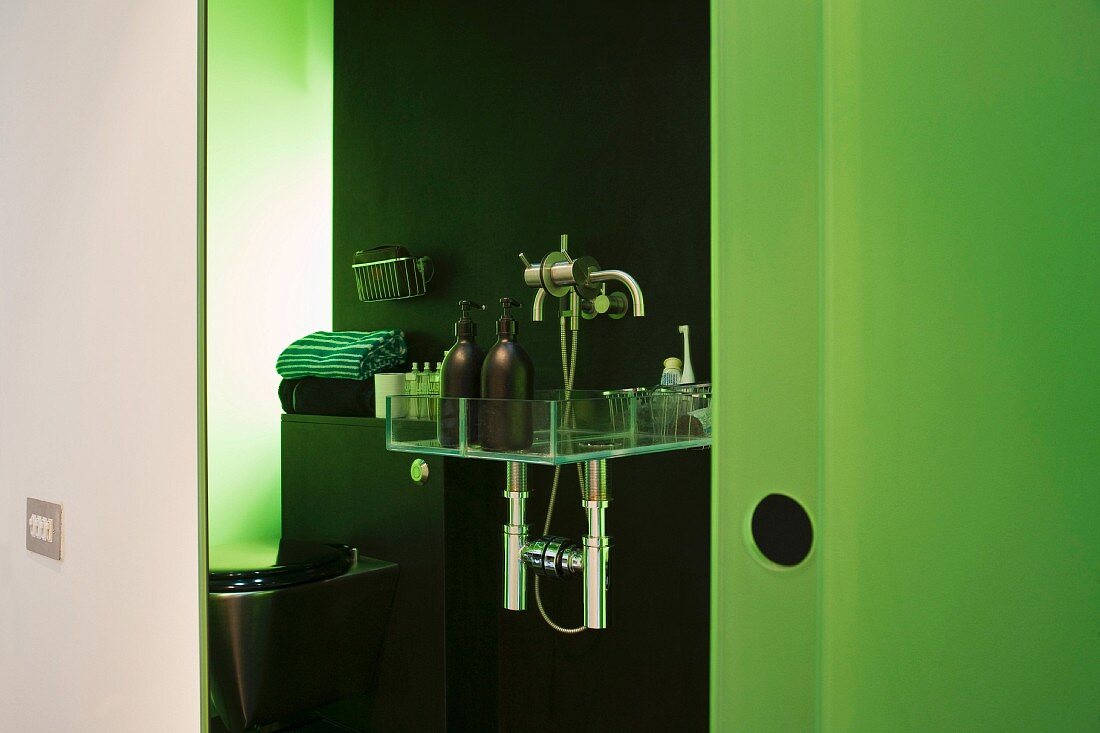 A view into an open-plan bathroom with bathing utensils on a shelf and a green glass wall