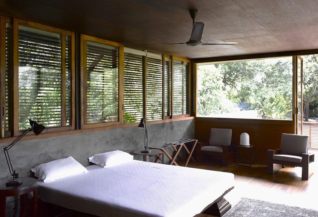Double bed with white bed linen in front of half-height concrete wall and wooden blinds on windows