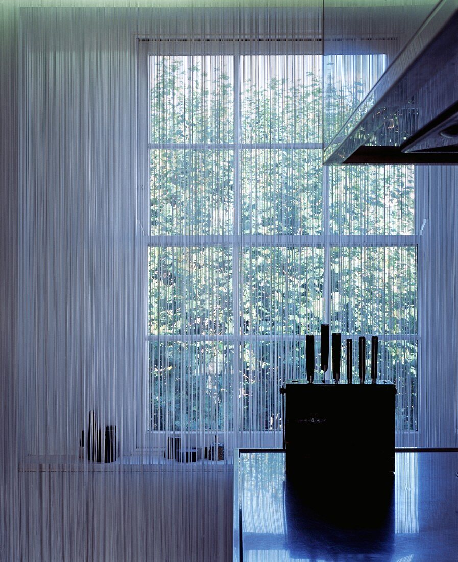 Spiegelung eines transparenten Schnurvorhangs in den Flächen einer Edelstahlküche und Messerblock im Gegenlicht