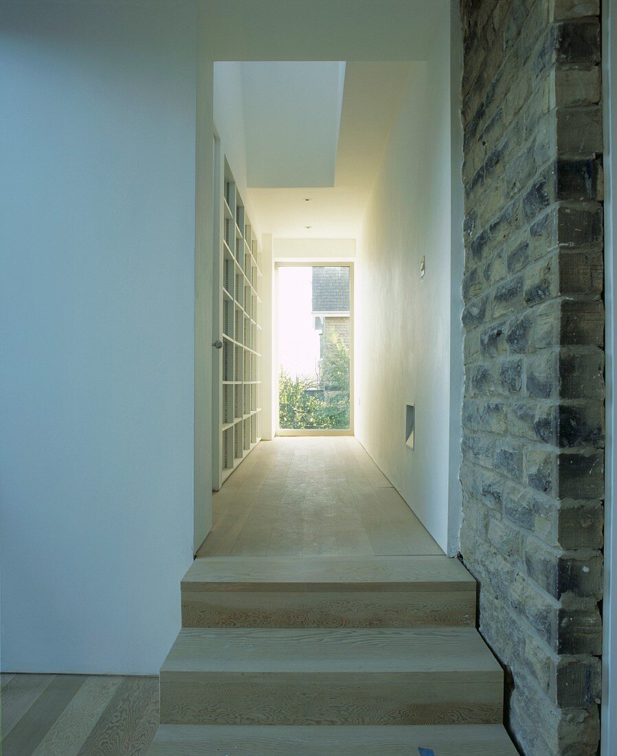 Holzstufen mit begleitender Natursteinwand zu schmalem Regalraum mit Glastür am Ende und Galerieöffnung nach oben