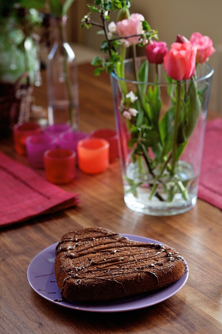 A heart-shaped chocolate cake with flowers and candles in the background