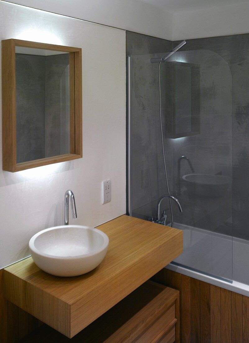 Modern washstand with white ceramic bowl on wooden pedestal and bathtub behind a glass partition
