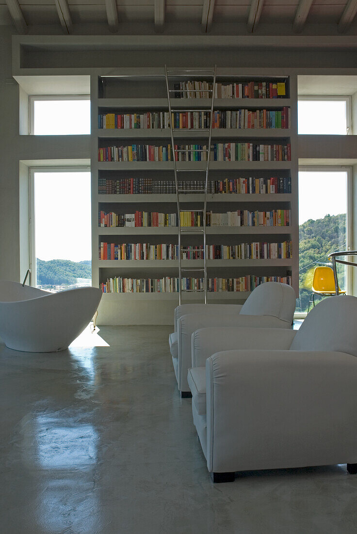 High shelf with books and white leather armchairs in minimalist living room with concrete floor