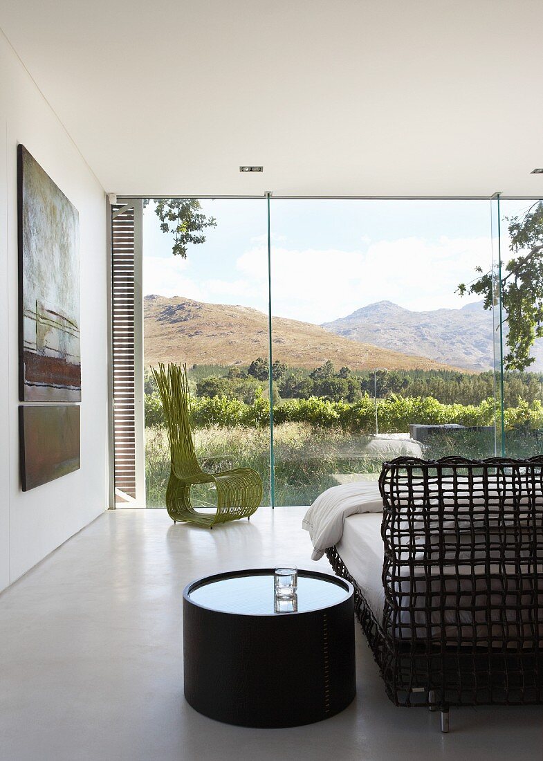 Bedroom with amazing view of landscape through frameless glass wall - round night stand next to delicate latticed bed frame