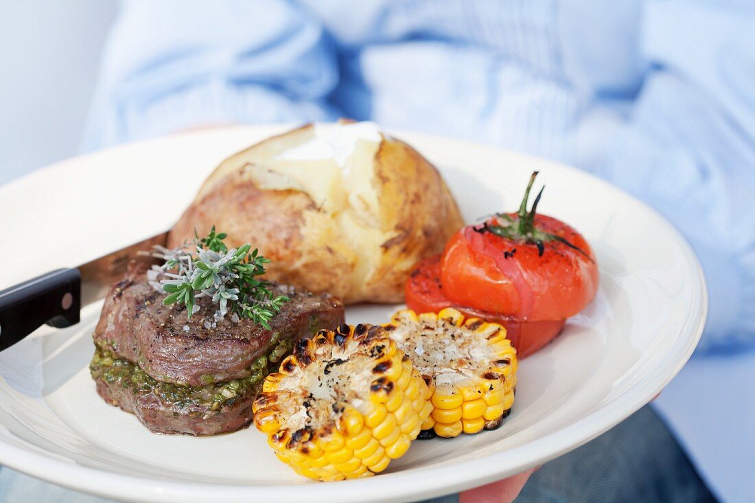 A person holding a plate of grilled beef medallions, a jacket potato and grilled tomatoes and corn on the cob