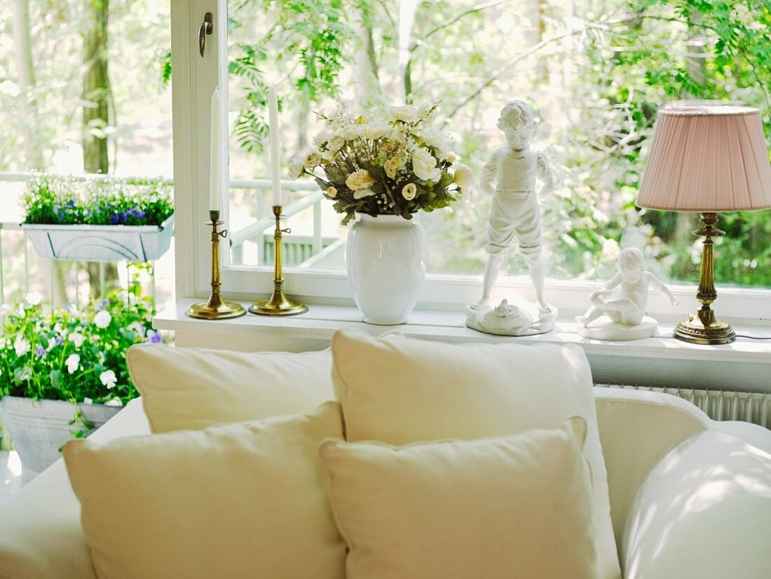 Cushions on white, upholstered armchair below large window decorated with flowers and vintage-style ornaments