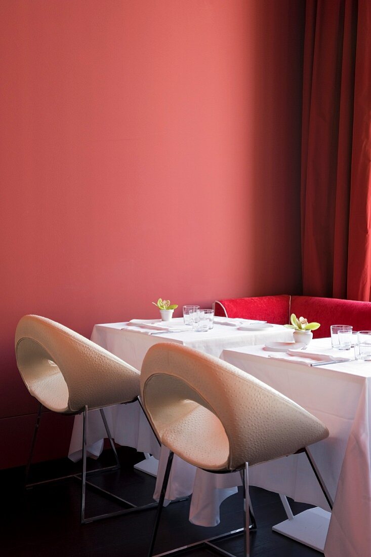 Set tables for two with white and deep pink designer chairs against brick-red wall