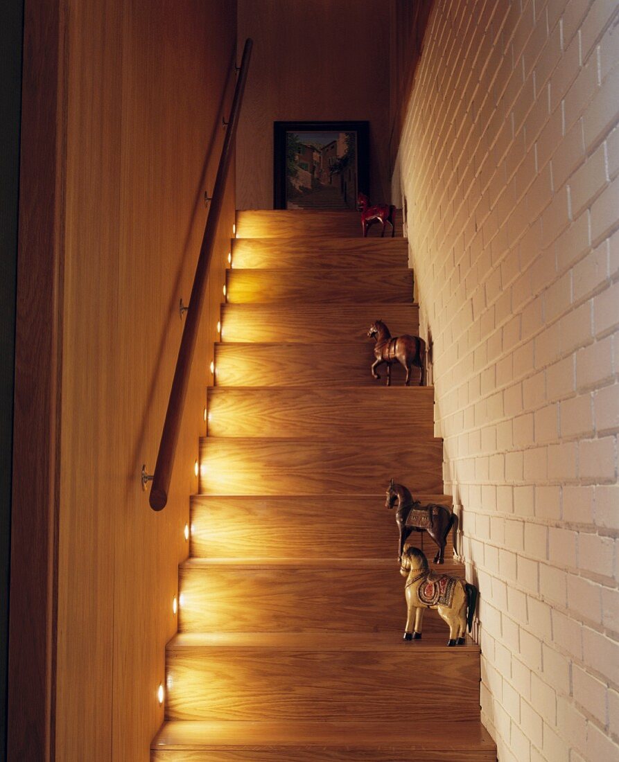 Collection of painted wooden horses on illuminated treads of staircase next to white-painted brick wall