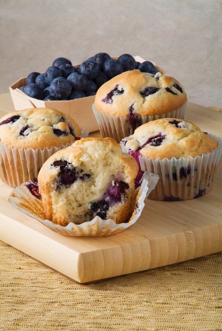 Four Blueberry Muffins on a Cutting Board with Fresh Blueberries; On Bitten