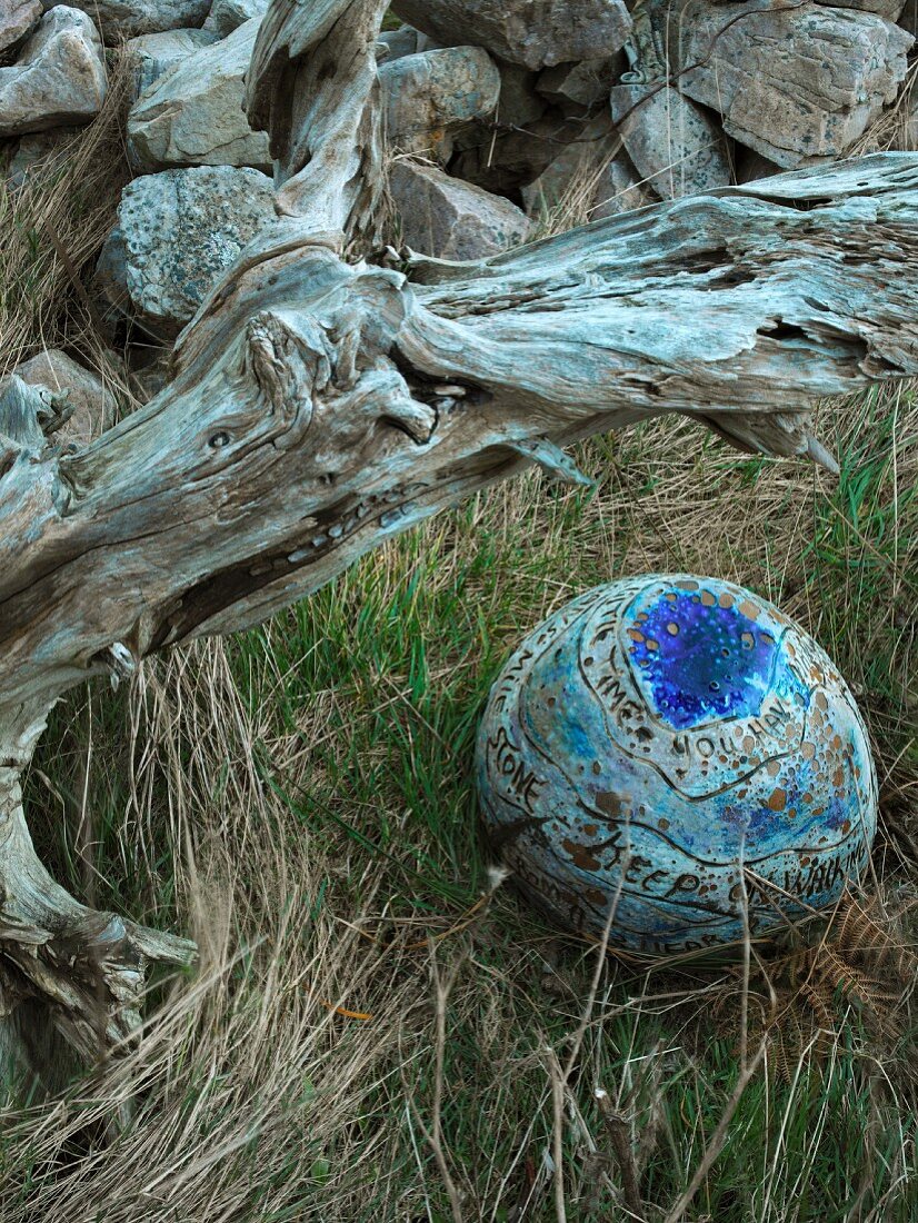 Blau glasierte Tonkugel mit Schriftband im Gras neben Wurzelholz