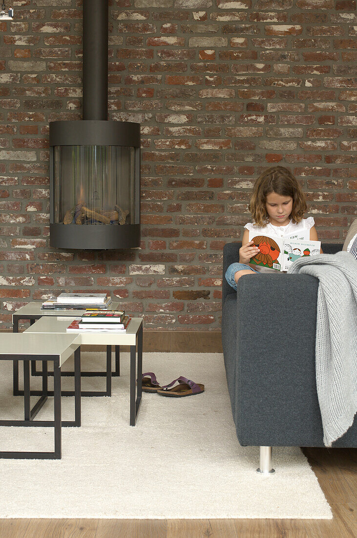 Girl reading on sofa, hanging fireplace in front of brick wall and coffee tables