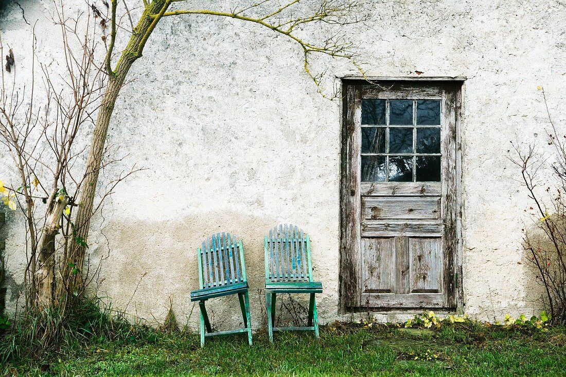 Two wooden chairs against exterior wall next to weathered door