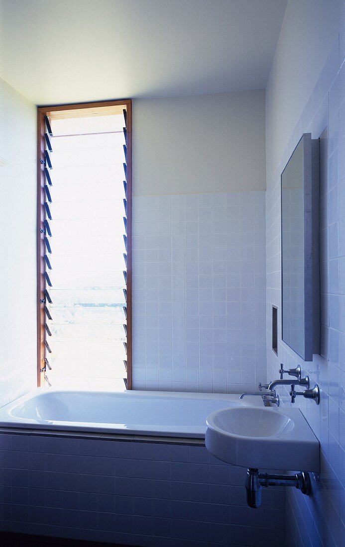 Corner of modern, white-tiled bathroom with bathtub below window with tilted glass slats