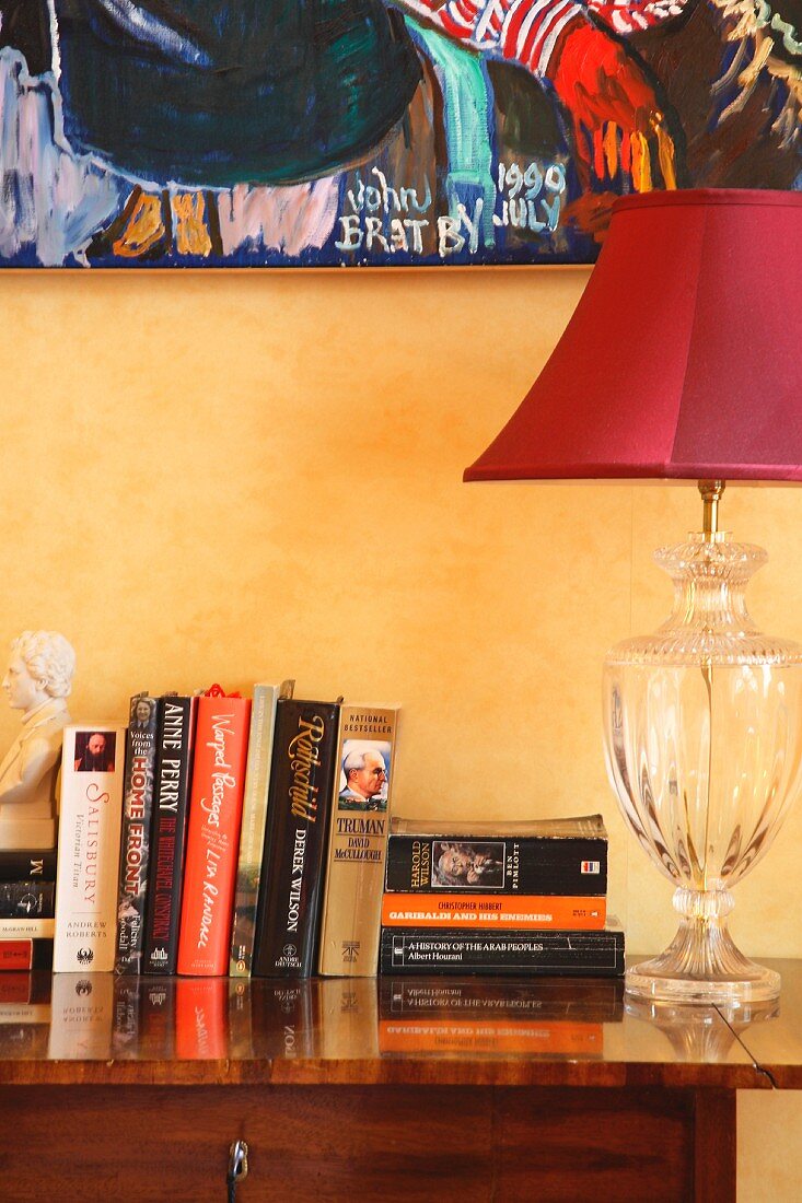 Bücher neben roter Lampe mit verziertem Glasfuss auf antiker Kommode