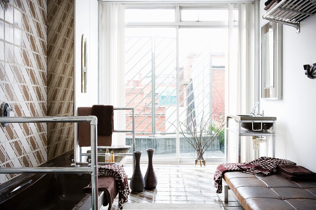 Blick durch raumhohe Fensterfront auf städtische Dachlandschaft in unkonventionellem Badezimmer mit Edelstahlgestellen und Lederliege
