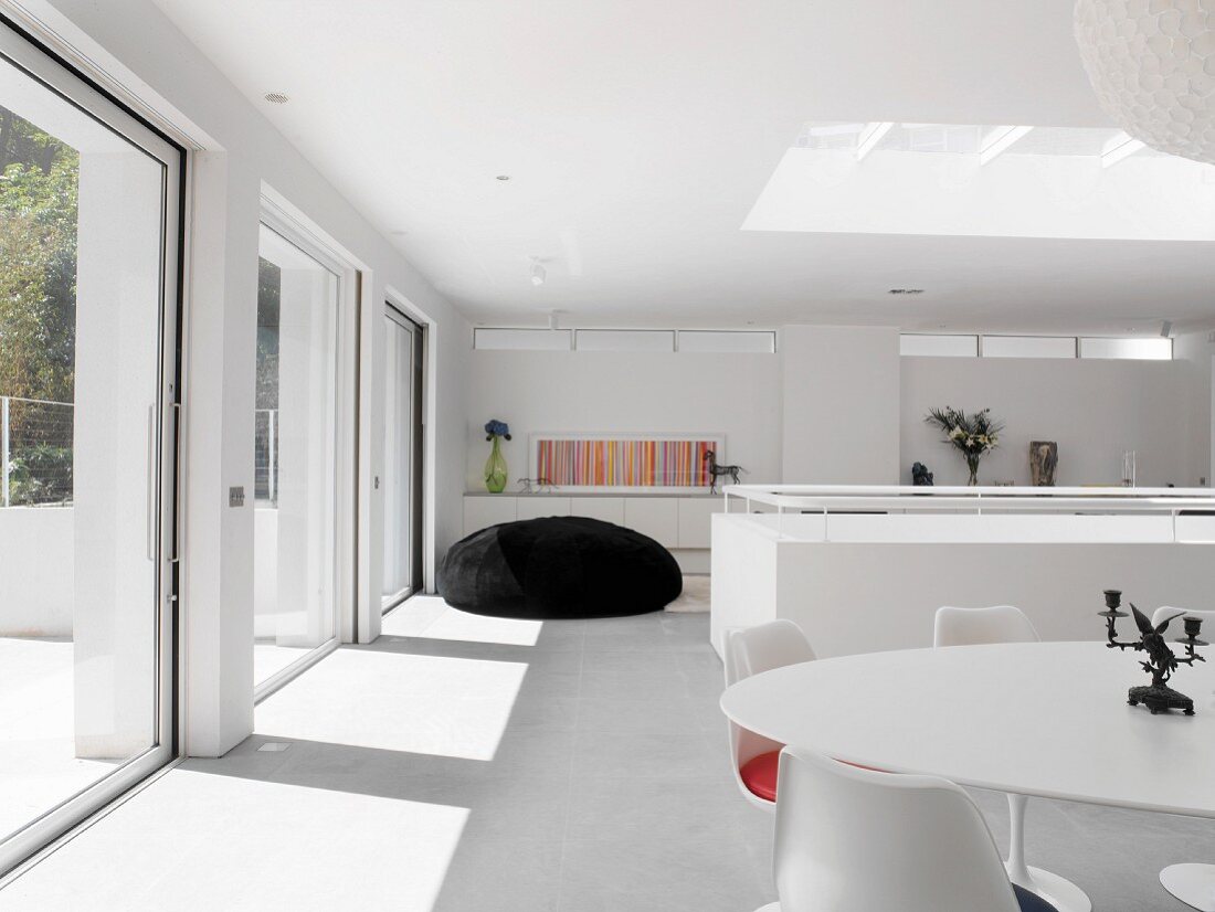 Dining area with classic designer pieces and oversized beanbag in white, sunny room with skylight and long glass wall with sliding doors