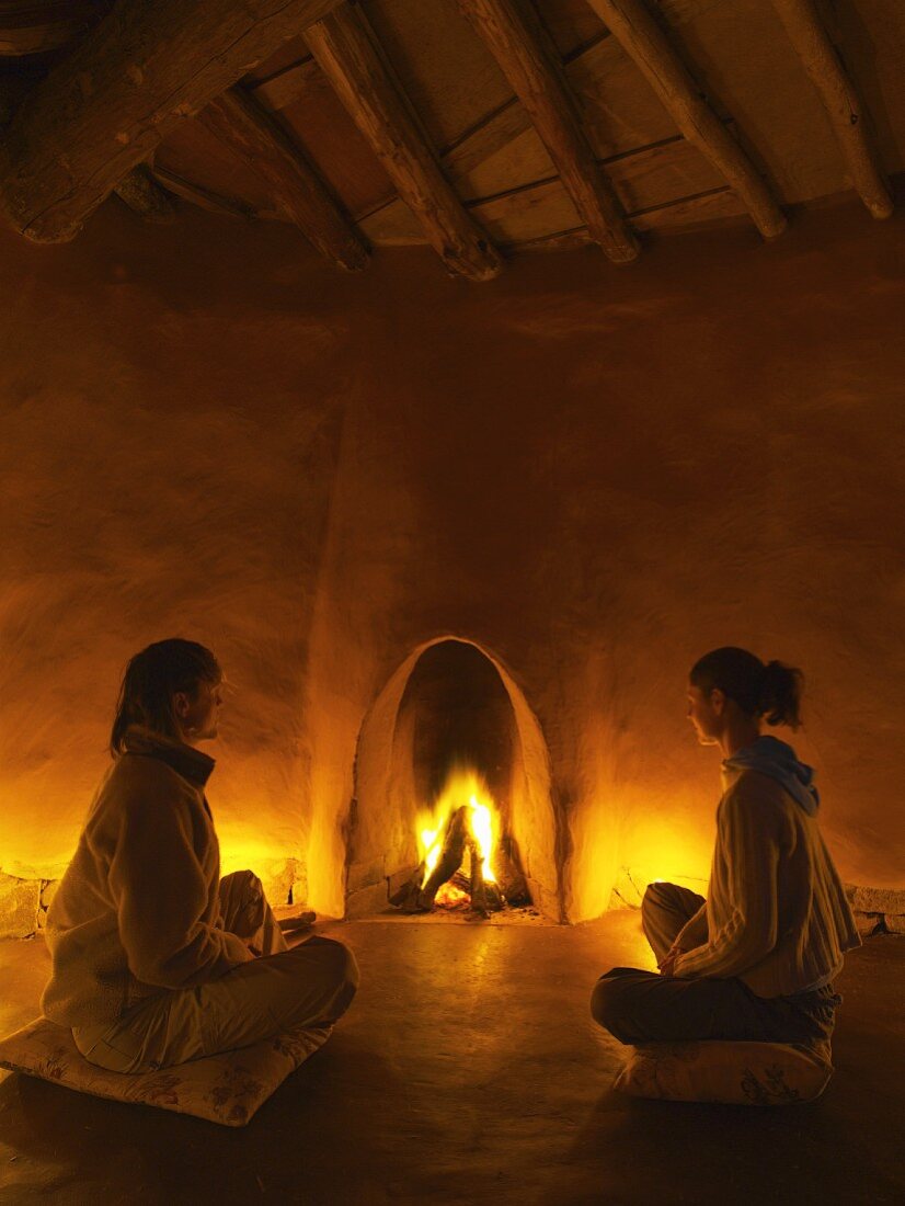 Meditative Stimmung in einfachem, organisch gestaltetem Lehmbau - zwei Frauen auf Sitzkissen vor loderndem Kaminfeuer