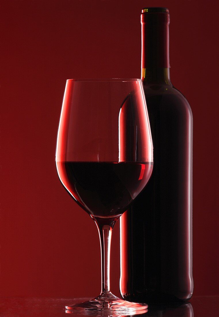 Rotweinglas und Rotweinflasche vor rotem Hintergrund