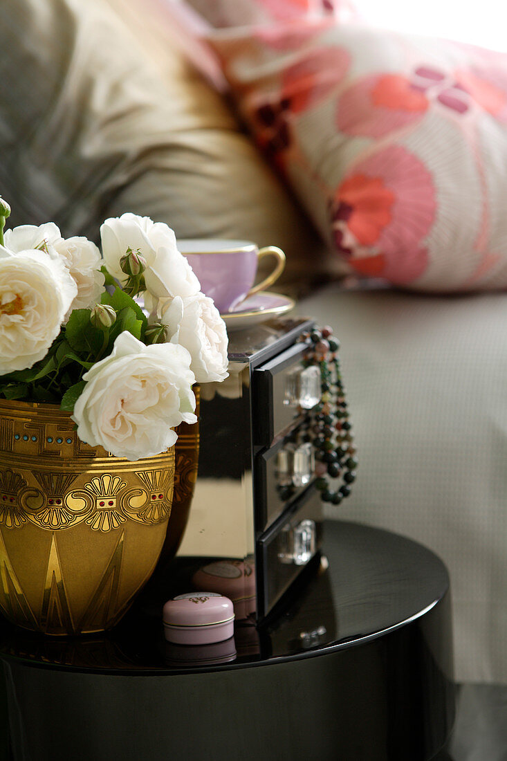 White roses in brass vase next to box with jewellery hanging of of drawers on black side table