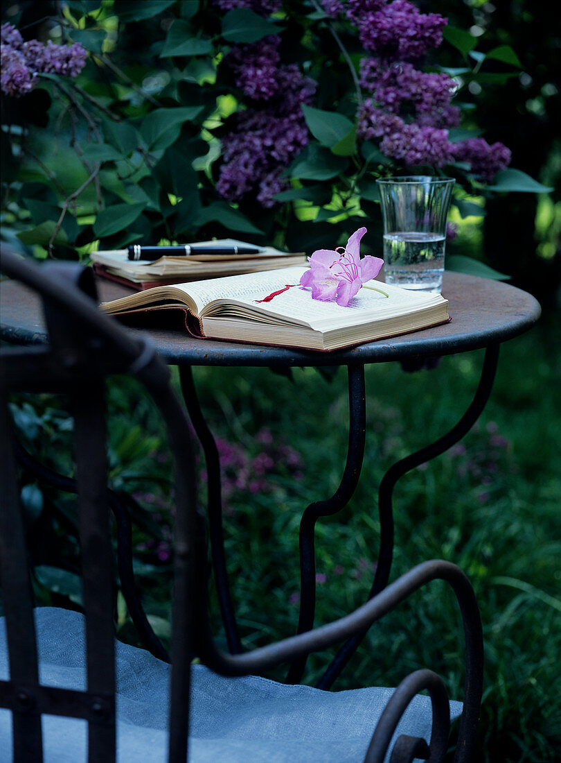 Aufgeschlagenes Buch auf Gartentisch aus Metall vor lila blühendem Flieder